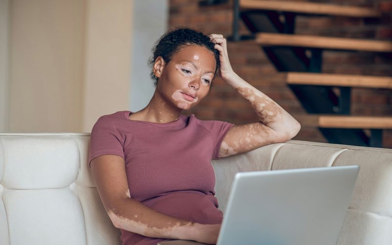 A person sits at a desk, smiling and resting their head on one hand, with shelves and books in the background.