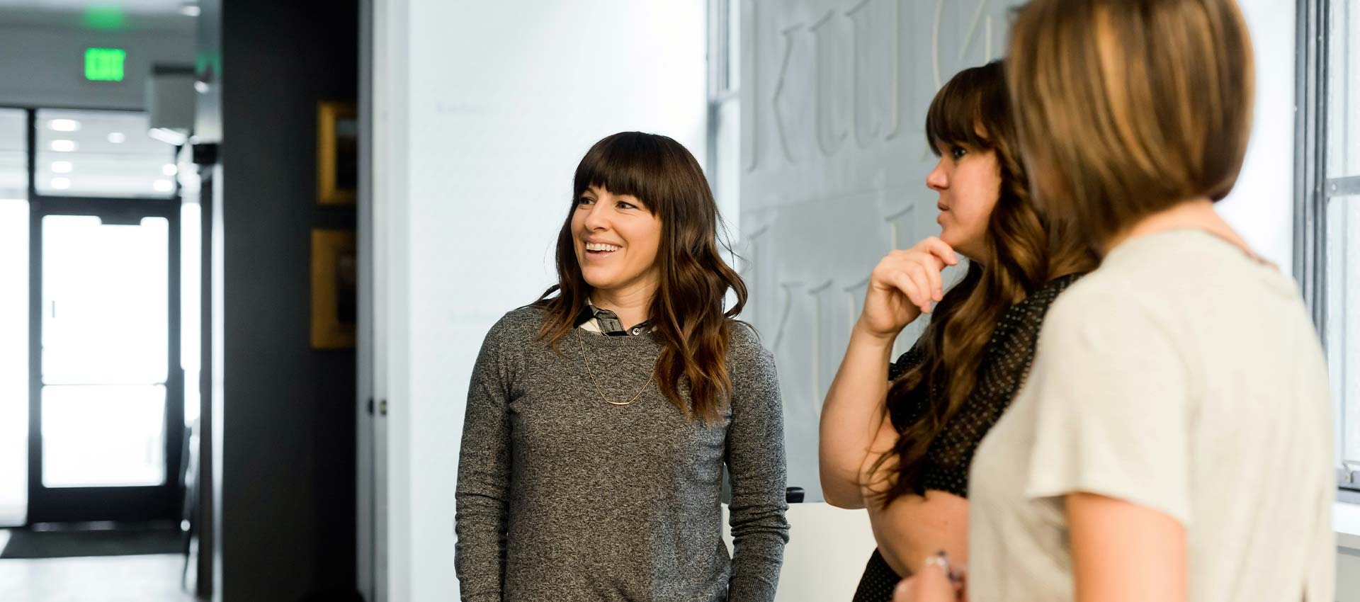 A person with long hair stands smiling, engaged in conversation with another individual. They are in a modern indoor setting with minimalistic decor and soft lighting.