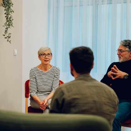 Three people are seated in a circle, engaged in conversation. A woman with glasses sits on the left, a man gestures with hands on the right, in a softly lit room.