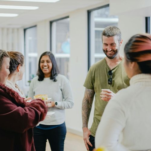 A group of five people stand in a bright room, engaging in friendly conversation. They are casually dressed and appear upbeat, with large windows in the background.