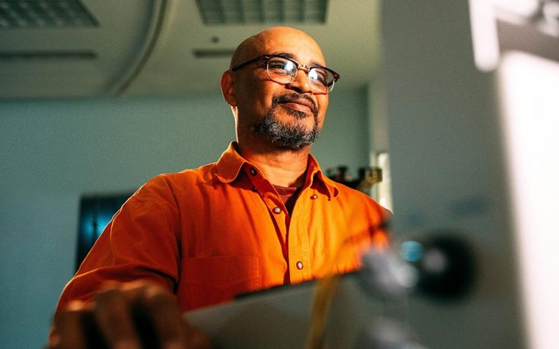 A person wearing an orange shirt is working at a computer in a well-lit office, focused on the screen with a slight smile.
