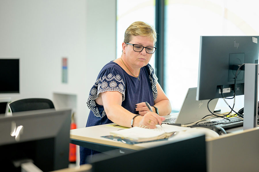 A person is sitting at a desk, writing in a notebook, with a laptop and a monitor in a bright, modern office environment.