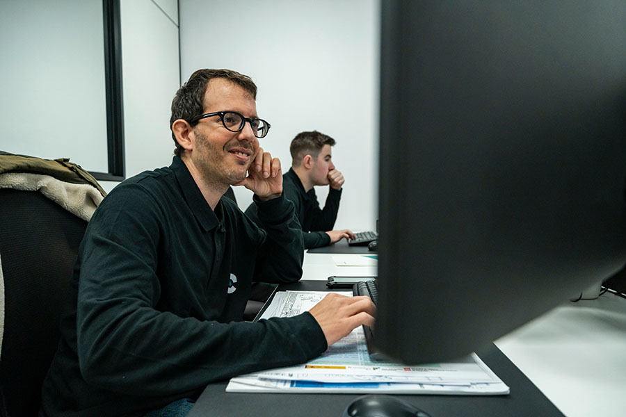 A man smiles while working at a computer in an office setting. Another person is visible in the background. The room is well-lit and has multiple desks and computers.