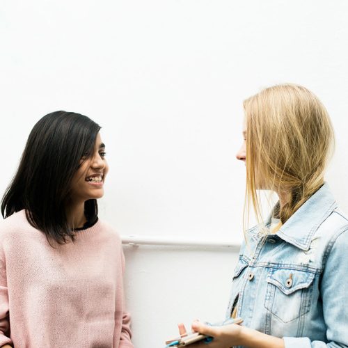 Two people engage in a friendly conversation indoors. One holds a clipboard and pen, while the other smiles. They stand against a plain white wall.