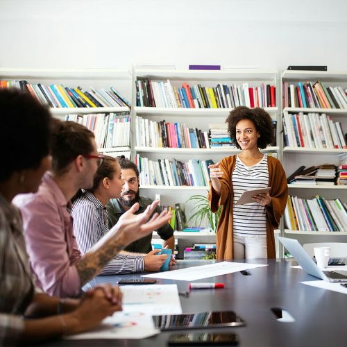 A woman holds a tablet, leading a discussion with four people seated at a table in a library. Bookshelves filled with colorful books create a vibrant backdrop.