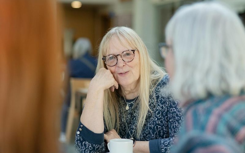A woman with long blonde hair and glasses sits at a table, resting her chin on her hand, engaged in conversation with others in a bright, indoor setting.