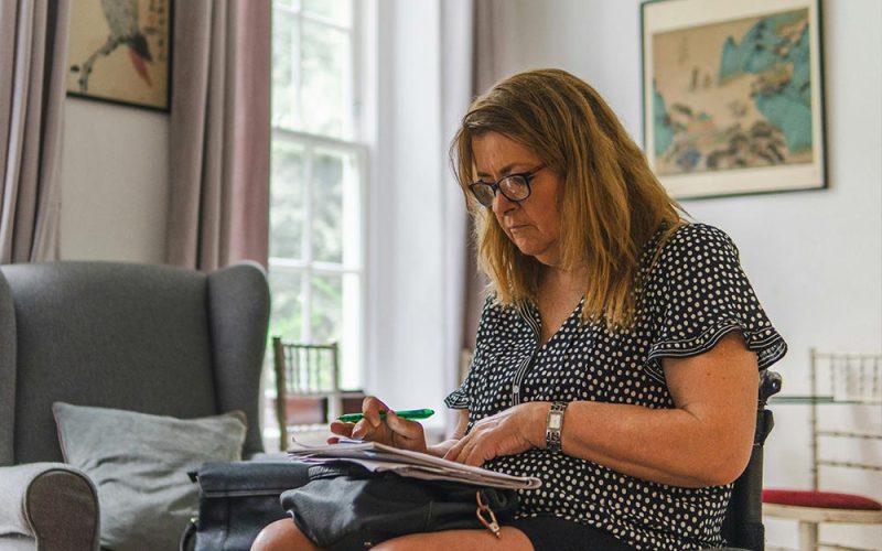 A woman with glasses writes on a notepad with a pen, seated in a comfortable, well-lit room with framed artwork and a cushioned chair nearby.