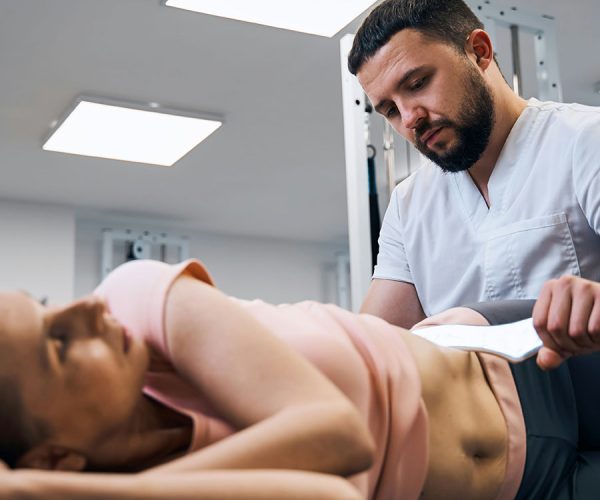A healthcare professional applies a therapeutic device to a patient's abdomen as she lies on her side in a clinical setting with bright overhead lighting.