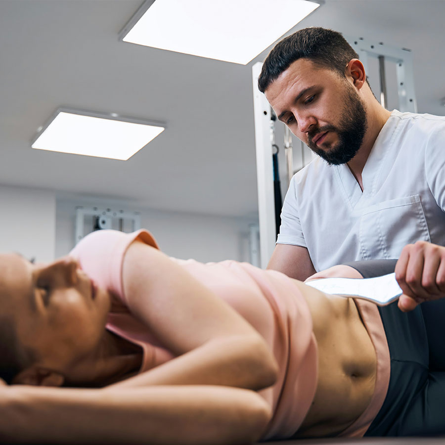 A healthcare professional applies a therapeutic device to a patient's abdomen as she lies on her side in a clinical setting with bright overhead lighting.
