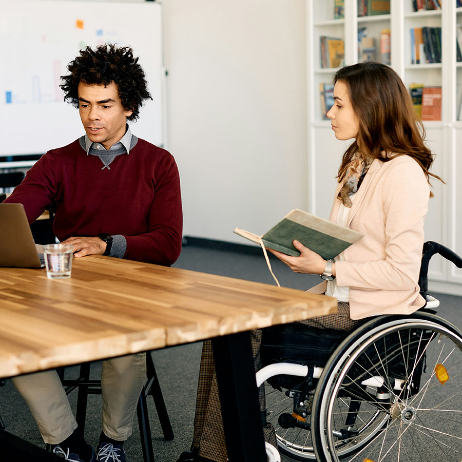 Man working on a laptop discusses with a woman in a wheelchair holding a notebook. They sit at a wooden table in an office with bookshelves.