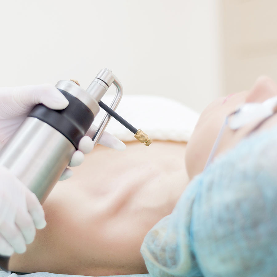 A medical professional holds a cryotherapy device, applying it to a patient's skin. The patient is lying on a treatment table, wearing protective goggles and a cap, in a clinical setting.