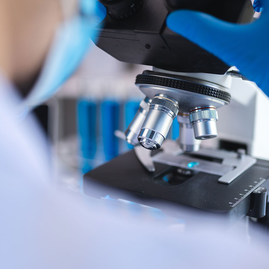 A scientist in blue gloves examines a sample through a microscope in a laboratory, with test tubes blurred in the background.