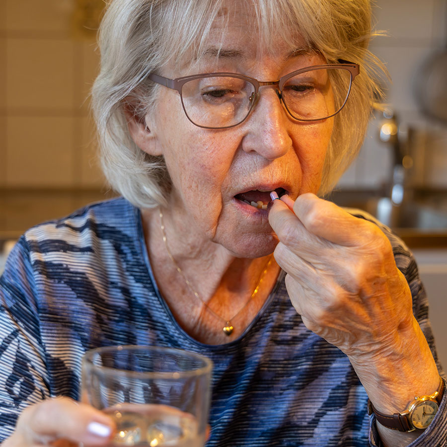 Elderly woman taking a pill with water, seated in a kitchen. She wears glasses and a striped shirt, focusing on the action of swallowing medication.