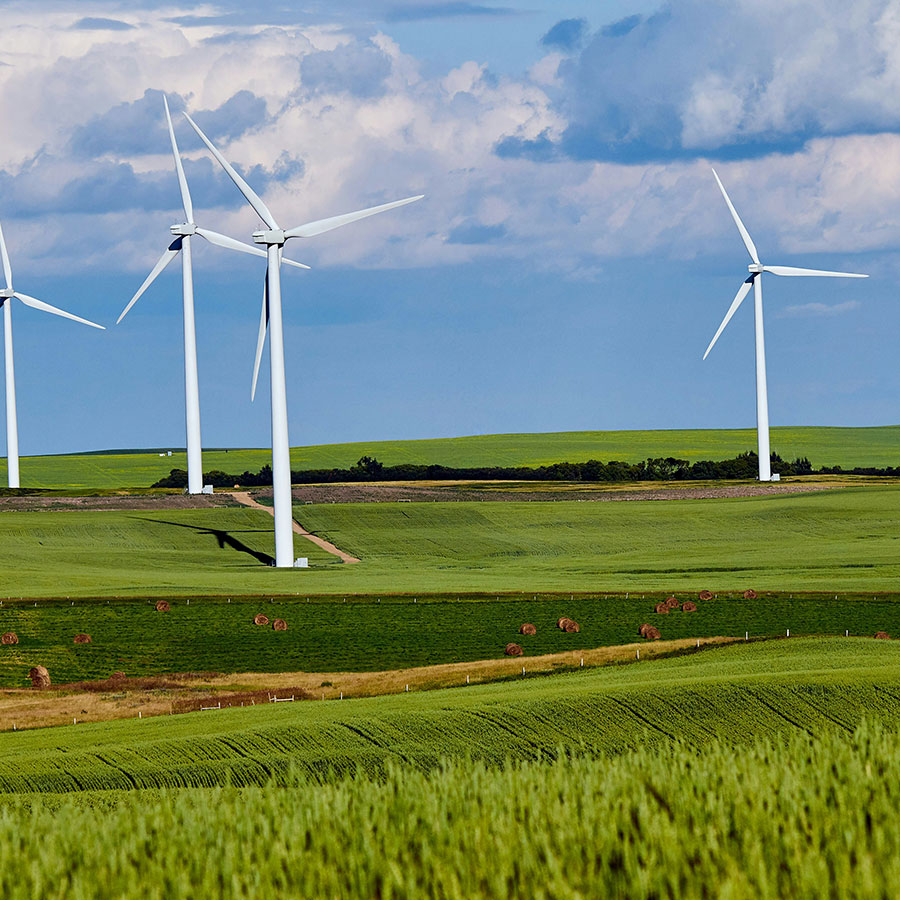 Wind turbines stand motionless on a vast, rolling green landscape under a cloudy blue sky. The scene conveys a sense of tranquility and sustainable energy in a rural setting.