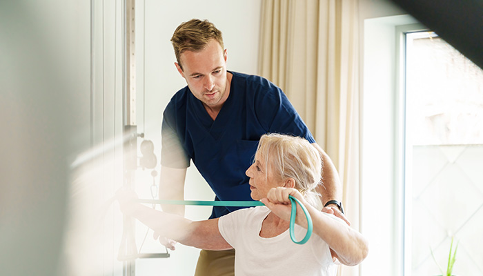 A healthcare professional assists an elderly woman with a green resistance band for exercise in a bright, well-lit room with cream curtains and large windows.