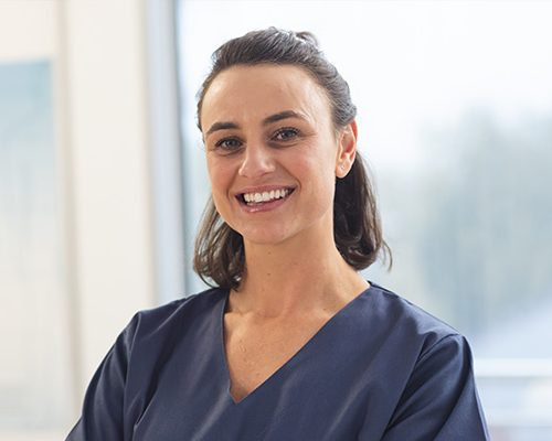 A person in a navy blue uniform smiles warmly, standing in a bright room with large windows, suggesting a healthcare environment.