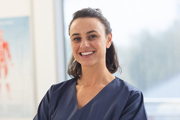 A person in a navy blue uniform smiles warmly, standing in a bright room with large windows, suggesting a healthcare environment.
