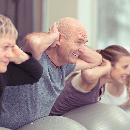 People exercise in a fitness class, leaning forward with hands behind their heads while balancing on stability balls. They are in a brightly lit room with large windows.