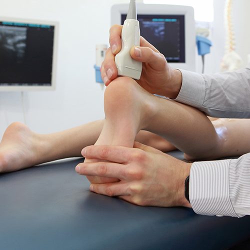 A healthcare professional uses an ultrasound device on a patient's ankle, examining it on a medical table. Background includes two monitors displaying medical images in a clinical setting.