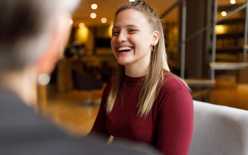 A person in a maroon top sits and smiles brightly, engaging in conversation with another person, in a warmly lit, modern indoor setting with blurred background elements.