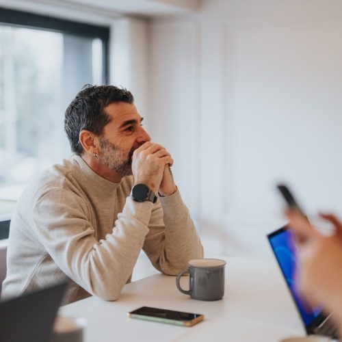 A person sits at a table, hands clasped, looking thoughtfully ahead. Nearby are a mug, smartphone, and open laptop in a bright, modern office setting.
