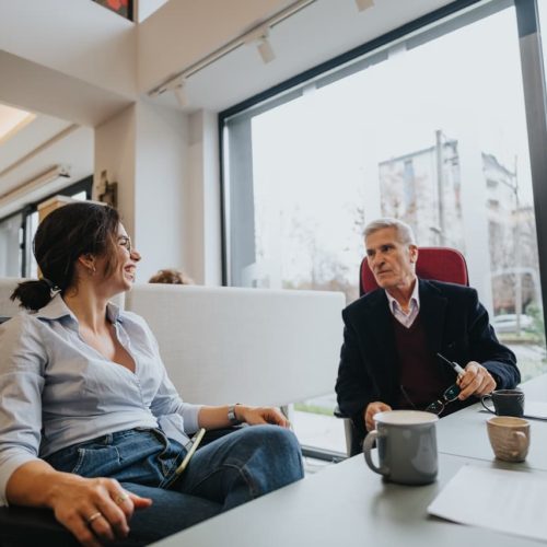 A women in a light blouse talks to a man in a suit as they sit at a desl together.