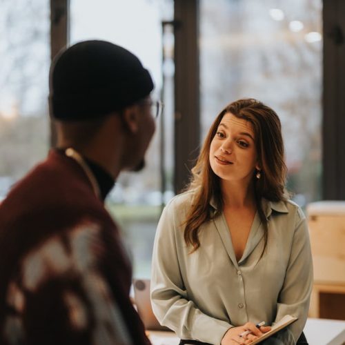 A woman, wearing a light blouse, listens and talks to a man in a beanie. They are in a modern, well-lit office with large windows in the background.