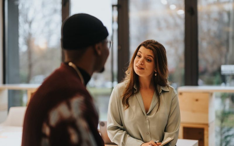 A woman, wearing a light blouse, listens and talks to a man in a beanie. They are in a modern, well-lit office with large windows in the background.