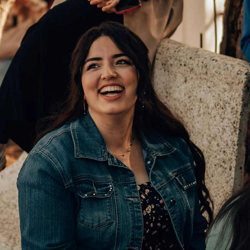 A woman wearing a denim jacket and a dress laughs while sat in a busy room.