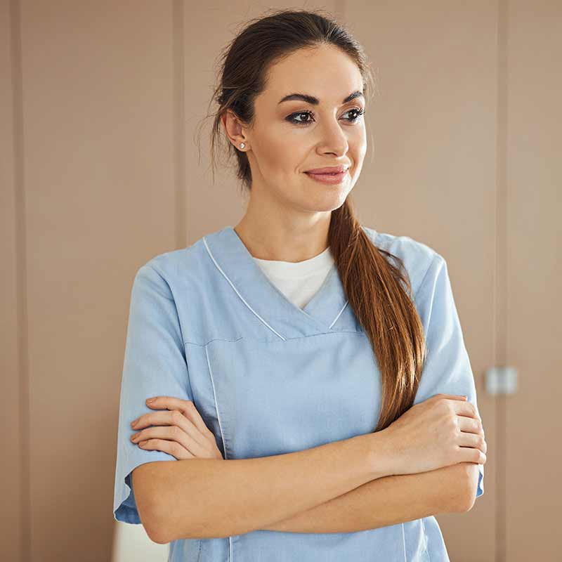 A woman wearing blue scrubs stands with her arms folded looking away from the camera.