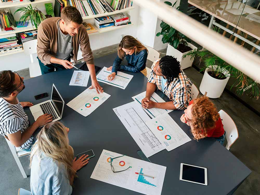 A group of people sit around a large table with laptops, tablets, and printed pieces of paper showing charts and graphs.