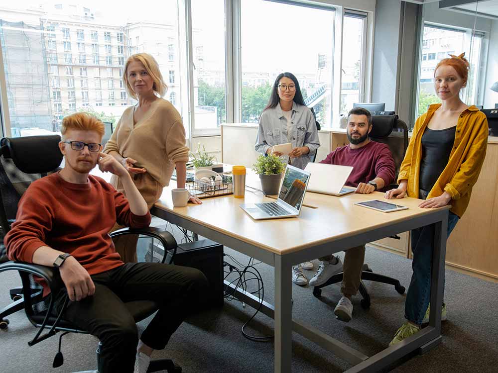 A group of men and women gather around a wooden office desk all looking directly at the camera.